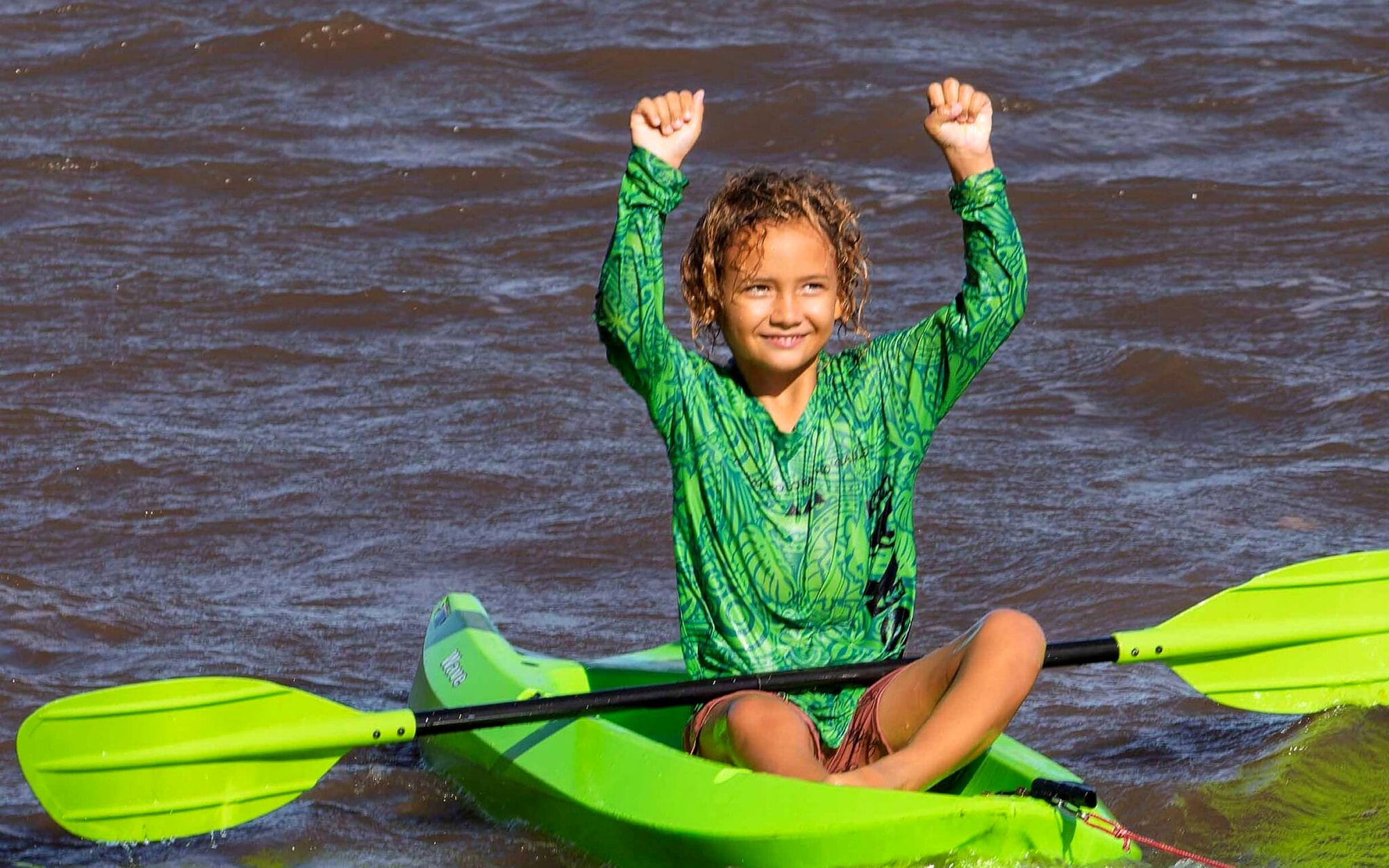 A young paddler cheers with both arms raised from a green kayak